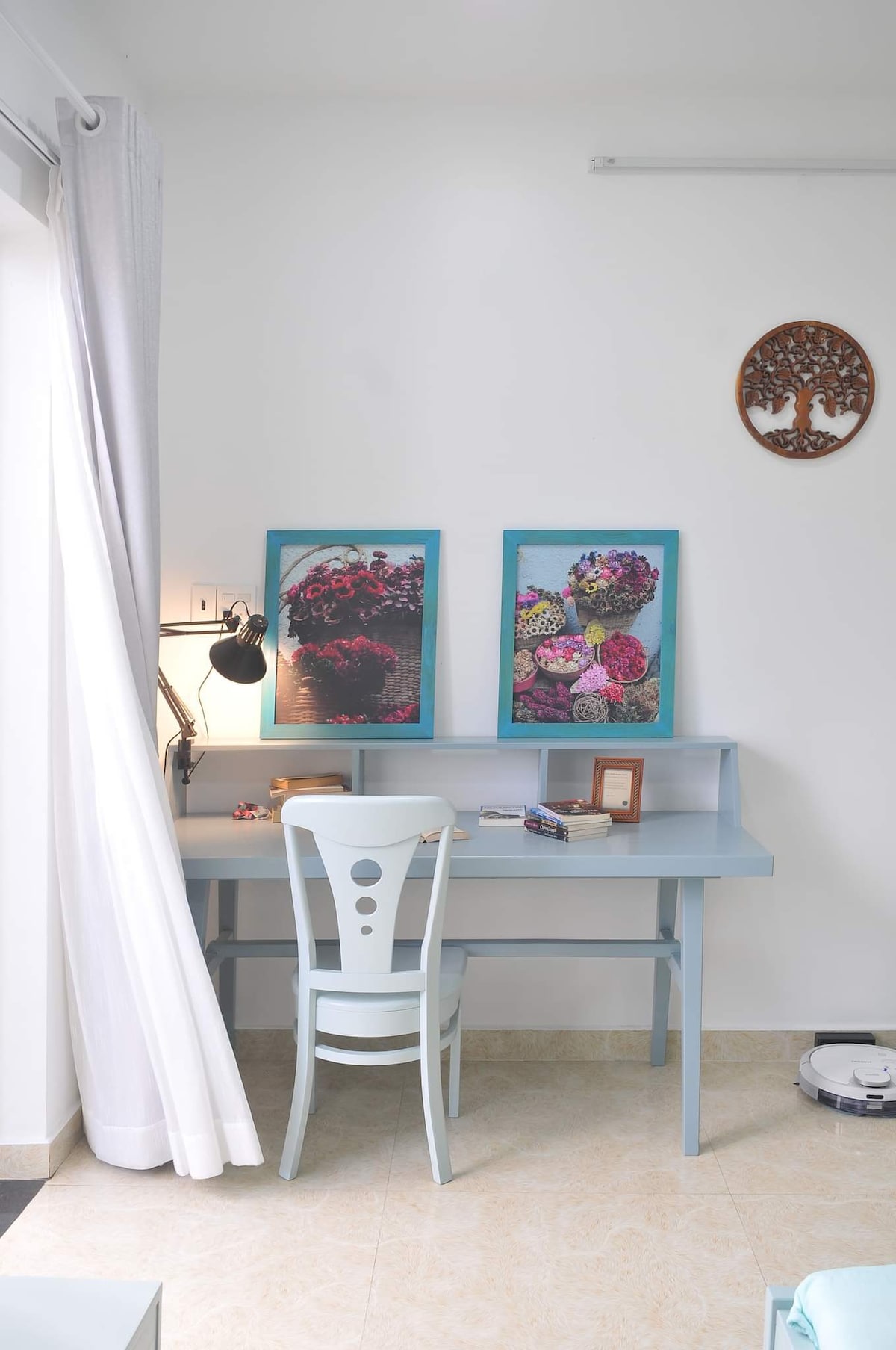A workspace is provided featuring a light blue desk with a matching chair, accompanied by two framed floral art pieces on the wall. A lamp illuminates the desk area, while light drapes softly hang beside a window, allowing natural light to filter into the room.