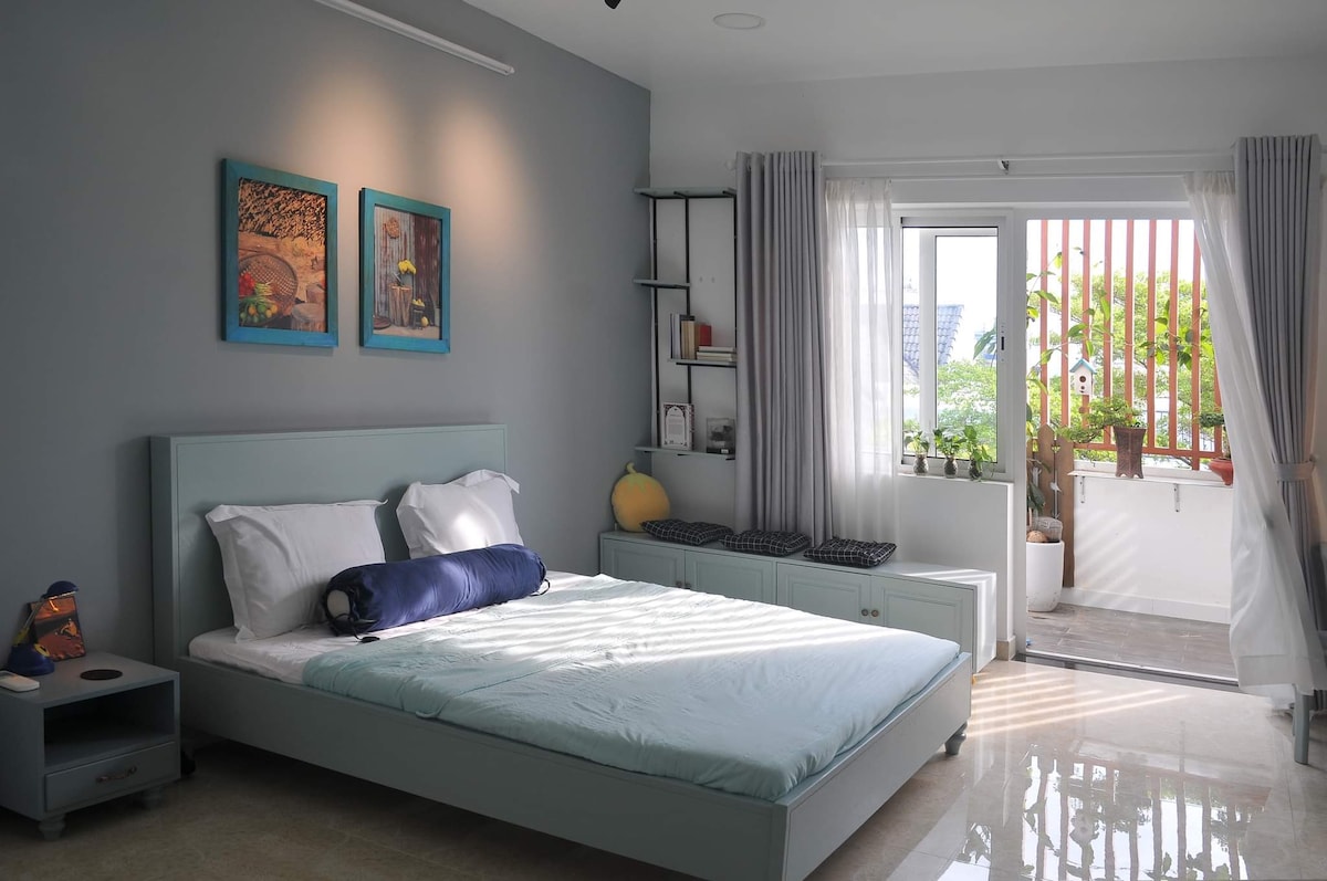 A spacious bedroom is filled with natural light, featuring a light blue bed with white linens and a large decorative pillow. Two framed artworks adorn the gray wall. A balcony is visible through the glass doors, which has potted plants and seating.