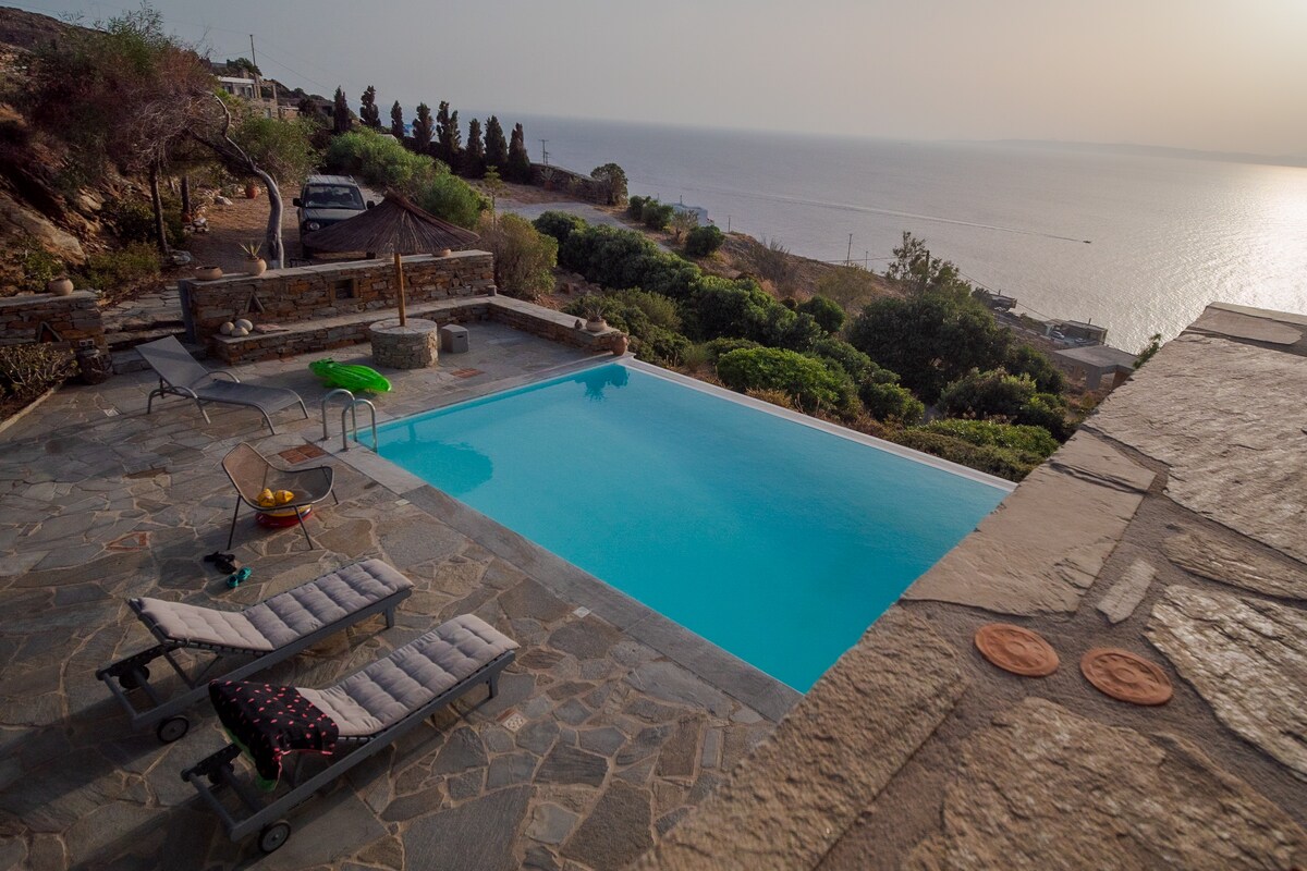 An outdoor area features a sparkling swimming pool surrounded by stone tiles. Sun loungers are positioned nearby, facing tranquil views of the Aegean Sea. Lush greenery is visible in the background, along with a distant coastline, under a warm, glowing sunset.