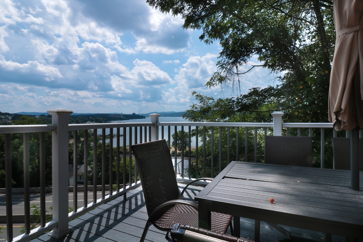 An outdoor deck area is featured with a wooden table and several brown wicker chairs. Lush greenery frames the scene, while expansive views of the Hudson River and cloudy sky create a serene backdrop.