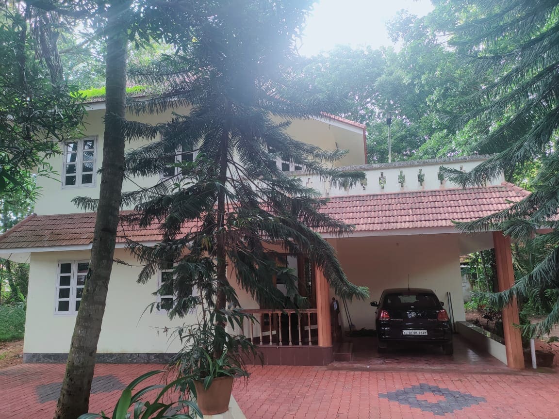 The house is set within a lush green landscape, featuring a front porch with a paved driveway and a parked car. Windows on the upper floor provide natural light, while palm and other trees frame the exterior, enhancing the serene environment.