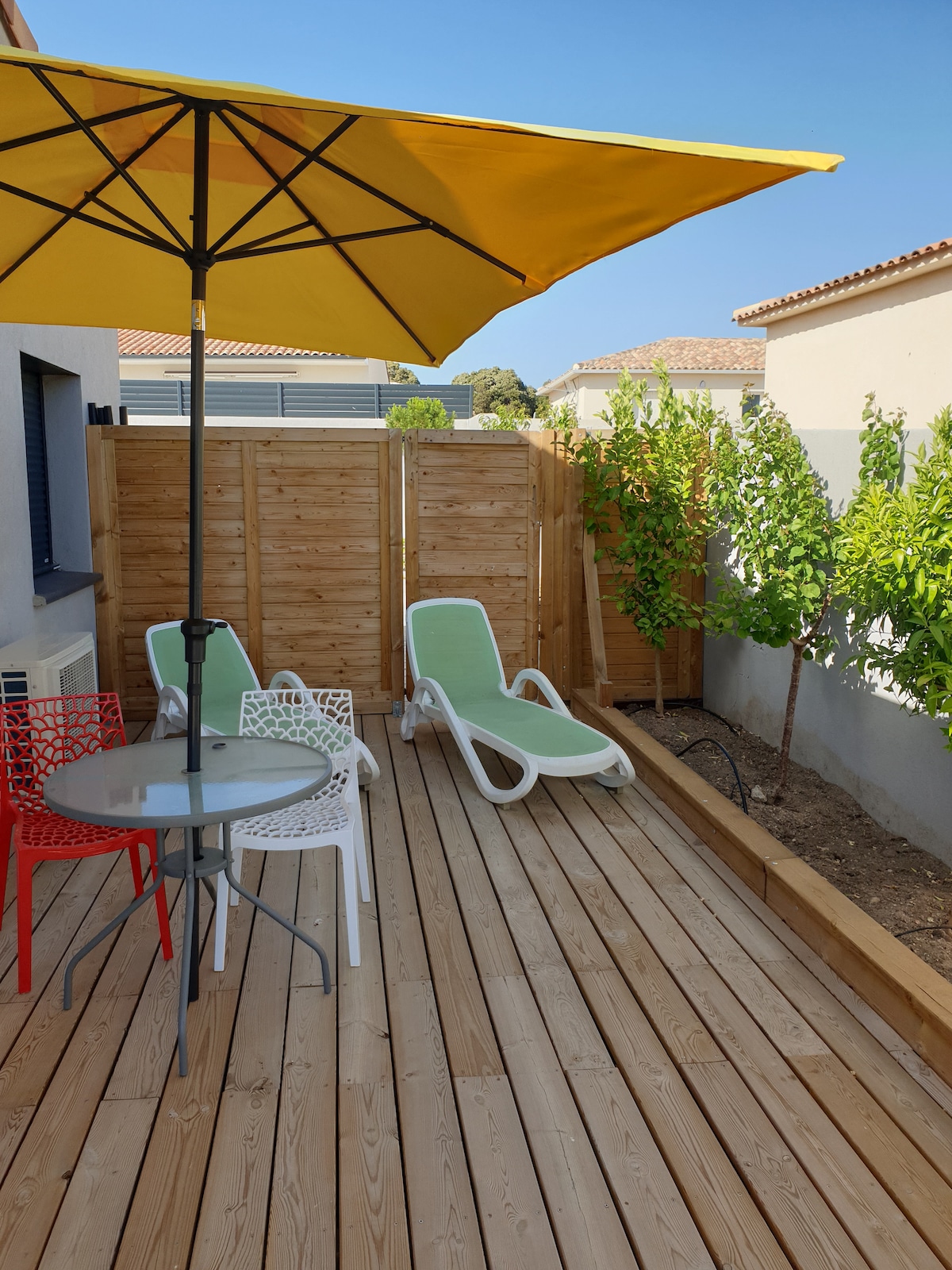 A private terrace is presented with a wooden deck, featuring a round table surrounded by four chairs. A large yellow umbrella provides shade over two loungers positioned for relaxation. Lush greenery is visible in the background, enhancing the outdoor space.