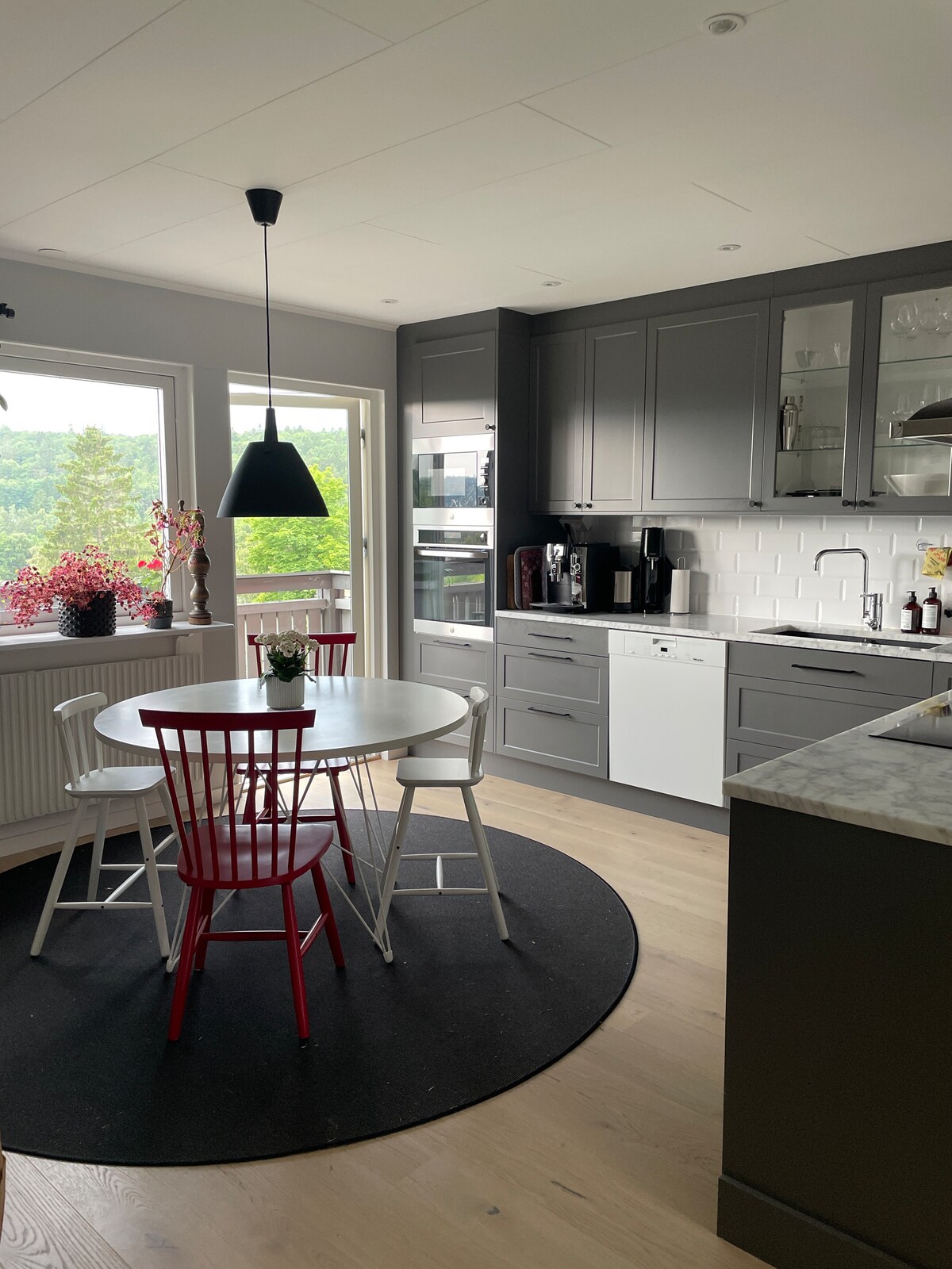 A modern kitchen features a circular dining table surrounded by three chairs, one in bright red. Dark cabinetry contrasts with a light countertop, while large windows allow natural light to fill the space, presenting views of greenery outside.