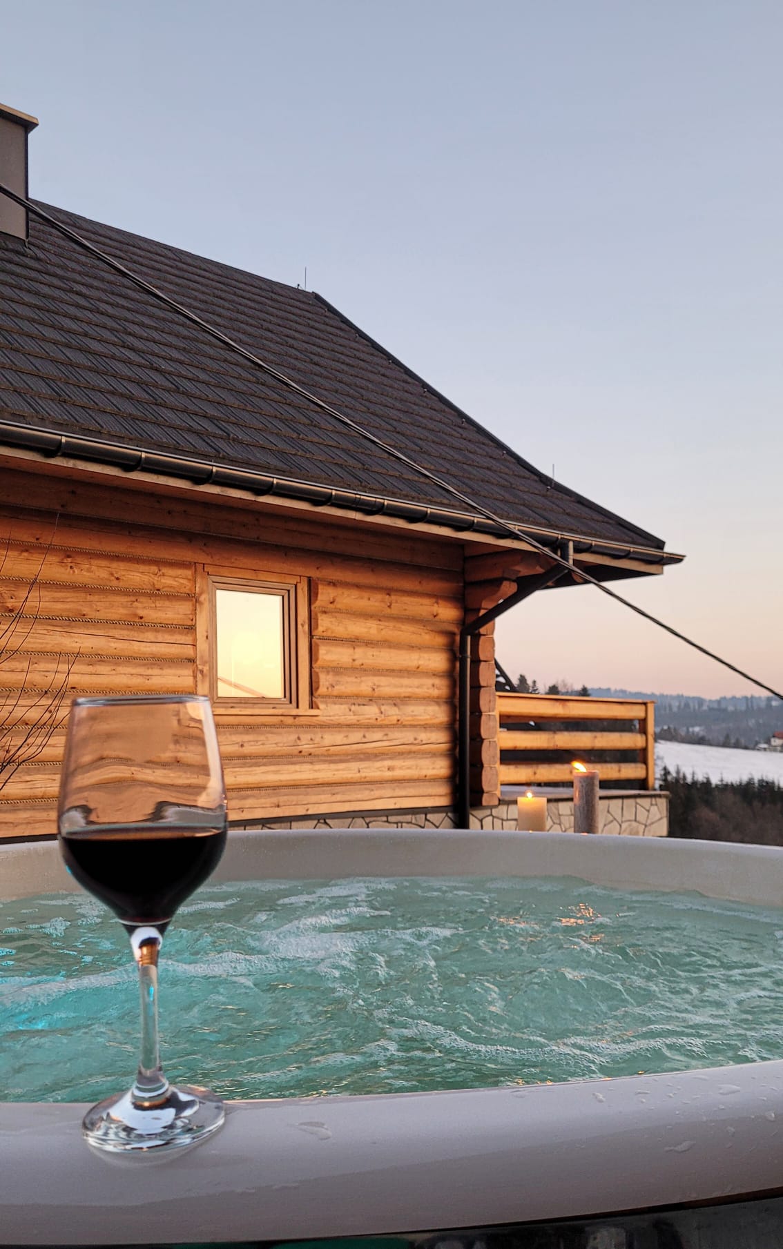An outdoor jacuzzi is visible in the foreground, with a glass of red wine placed on the edge. The wooden cabin exterior is shown, along with a view of the landscape and a soft sunset in the background.