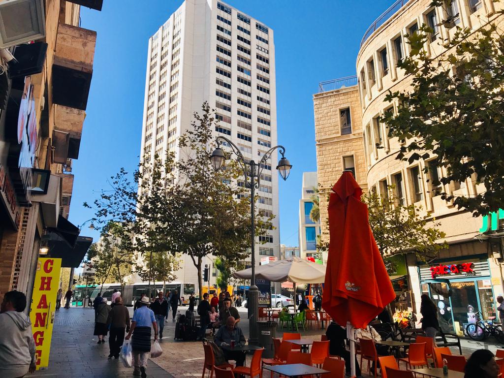 The image captures a bustling city street with vibrant orange umbrellas providing shade for outdoor seating. Modern buildings rise prominently against a clear blue sky, while pedestrians walk along the sidewalk. Trees line the street, adding greenery to the urban landscape.