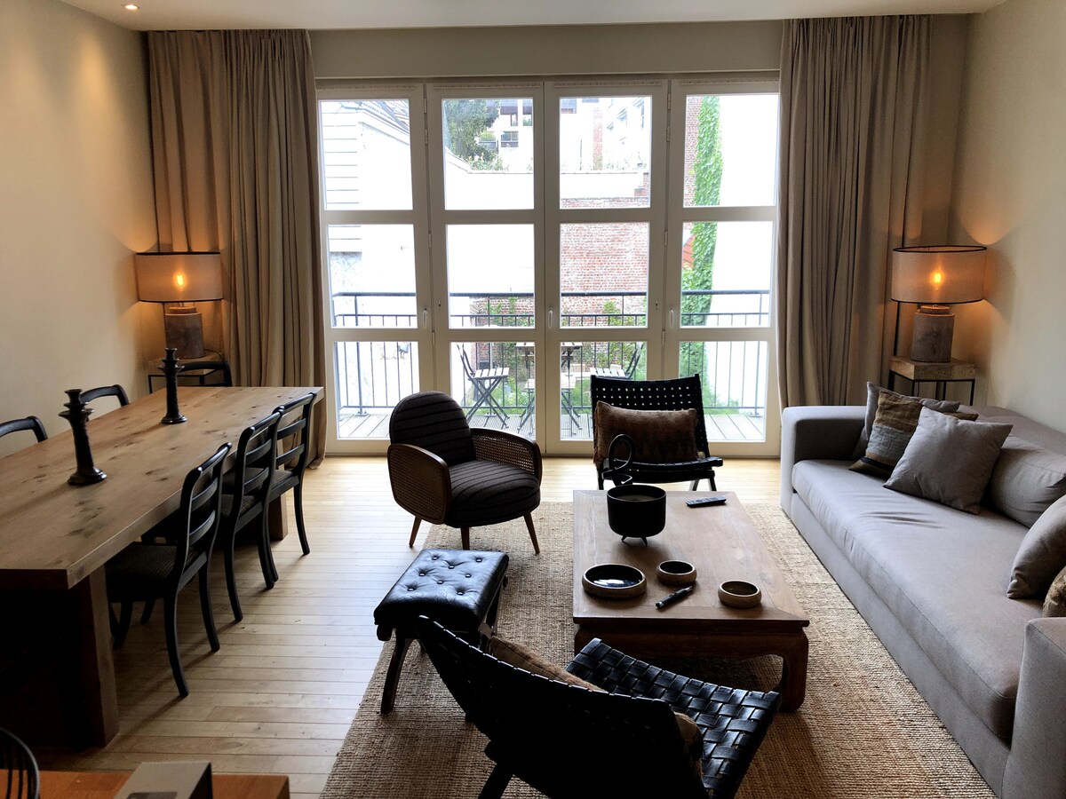 A spacious living area features large windows that provide natural light and views of the outdoors. A wooden dining table is surrounded by chairs, while a comfortable sofa and armchair invite relaxation. Textured rugs enhance the cozy feel of the room.