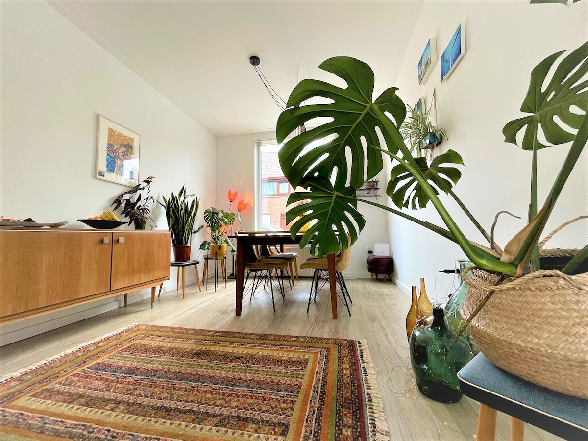 A light-filled dining area features a wooden dining table surrounded by chairs, offering space for six. A sideboard holds decorative plants and elegant vases. A colorful rug lies on the floor, adding texture to the room, while large plants bring a touch of nature indoors.