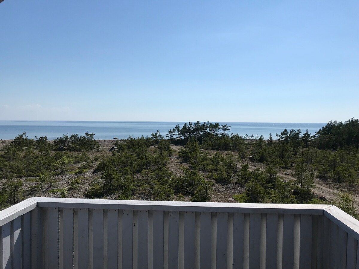 A panoramic view of the serene coastline is observed from the wooden balcony. The tranquil sea reflects the clear blue sky, while a scattering of trees line the shore. The peaceful natural landscape offers a calming backdrop.