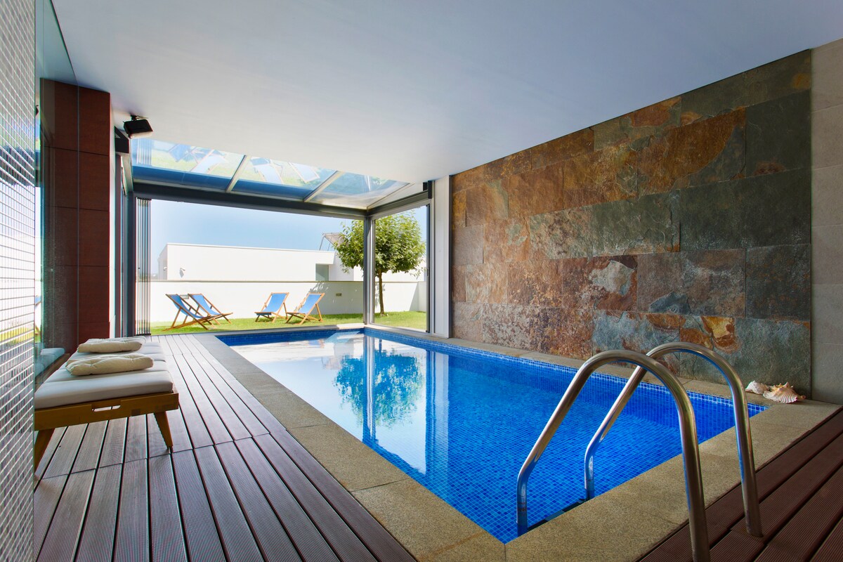 An indoor swimming pool is framed by large glass doors that open to a garden area. The pool is surrounded by wooden decking, with two lounge chairs placed nearby. A stone wall provides a natural accent to the modern space.