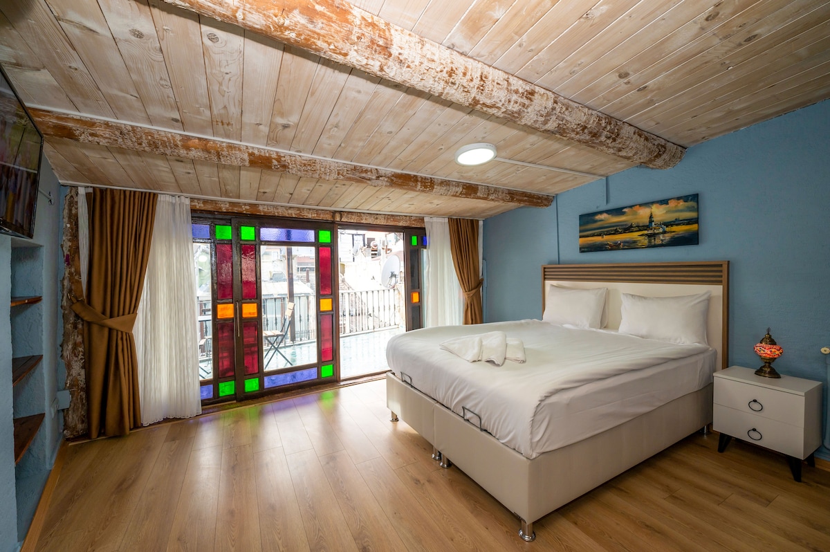A spacious bedroom features a large bed dressed in crisp white linens, complemented by decorative towels. Colorful stained glass windows provide vibrant light, while soft drapes frame the space. A small side table with an elegant lamp adds a touch of decor against the light blue walls.