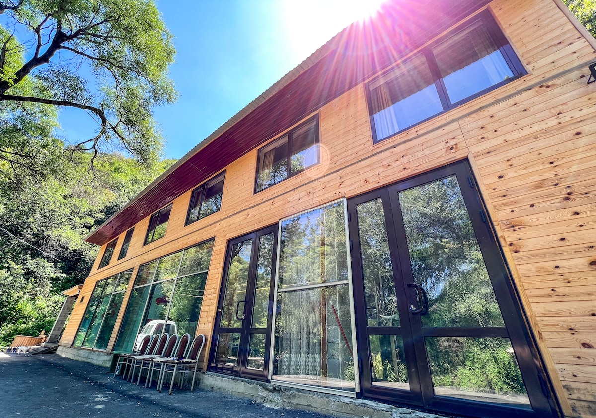 A wooden-clad building features large glass windows and doors that provide an expansive view of the outdoors. Sunlight reflects off the glass, illuminating the space. Several chairs are placed outside, enhancing the connection to the surrounding nature.