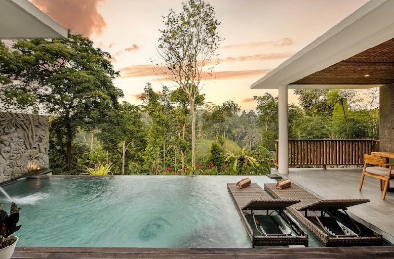 An outdoor swimming pool is surrounded by lush greenery, with a backdrop of trees and hills. Two lounge chairs are positioned beside the pool, and a covered terrace offers additional seating, creating a serene space to relax and enjoy the view.