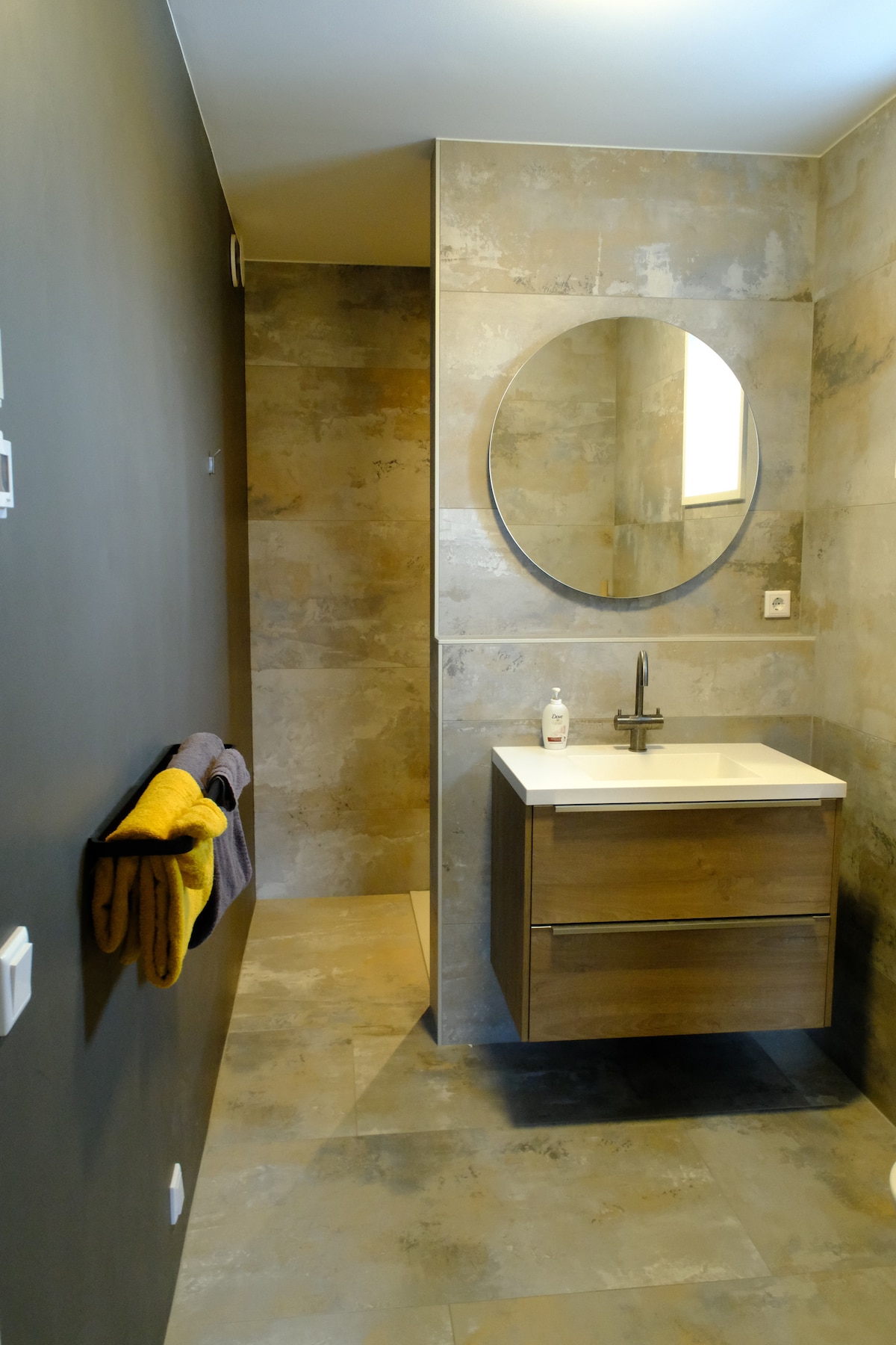 A modern bathroom features sleek stone tiles and a circular mirror above a minimalist vanity with a single basin. Soft towels in shades of purple and yellow are neatly hung beside the hand dryer. A spacious shower area is partially visible, contributing to the overall clean design.