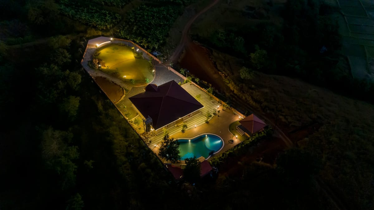 An aerial view captures a spacious villa surrounded by illuminated pathways and a tranquil swimming pool. The landscape features green foliage and open areas, highlighting the serene atmosphere of the property during nighttime. Soft lighting enhances the overall appeal of the villa's exterior.