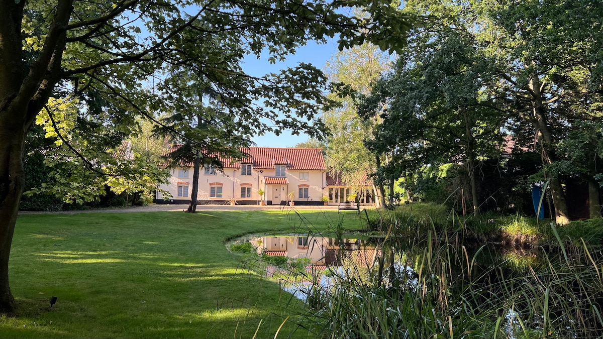 The tranquil grounds feature a lush green lawn bordered by trees, with a serene pond reflecting the exterior of the retreat house. Sunlight filters through the leaves, enhancing the peaceful ambiance of the countryside setting.