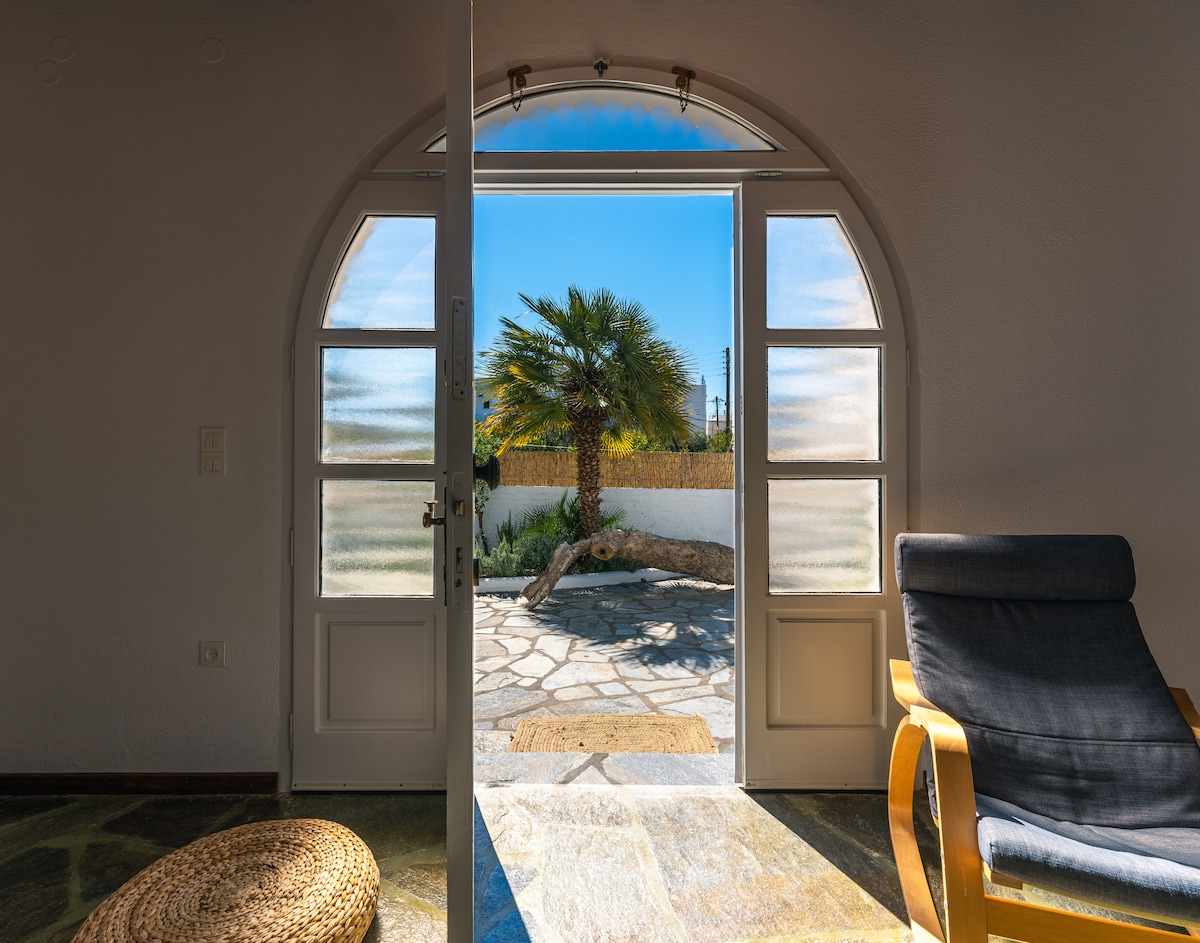 The entryway features arched doors that open to an outdoor deck, showcasing a palm tree and a well-maintained garden. Natural light streams into the interior, illuminating a comfortable chair and a woven rug, enhancing the seamless transition between indoor and outdoor spaces.