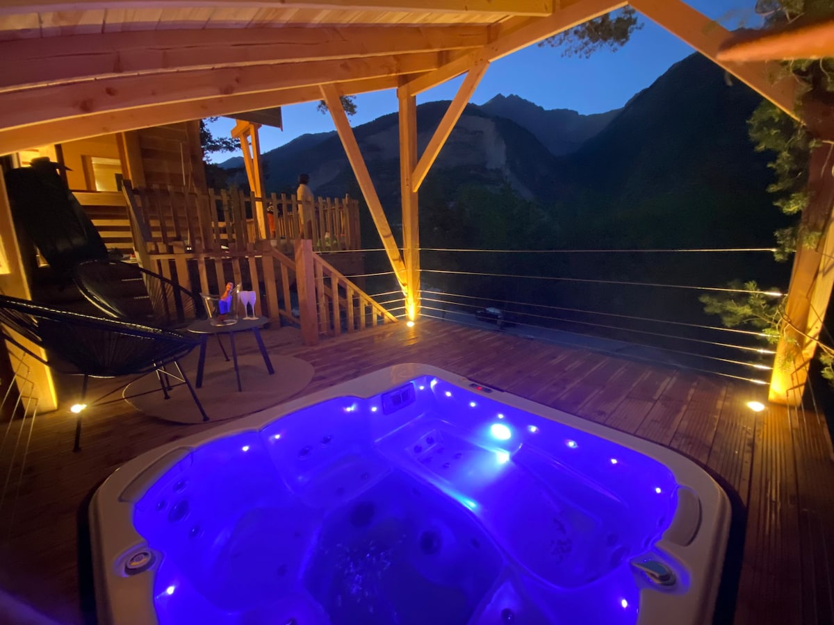 An outdoor spa area is illuminated by soft blue lights, surrounded by wooden decking. Two lounge chairs are positioned nearby, with scenic mountain views visible in the background. The tranquil atmosphere is enhanced by the evening setting, creating a peaceful retreat.