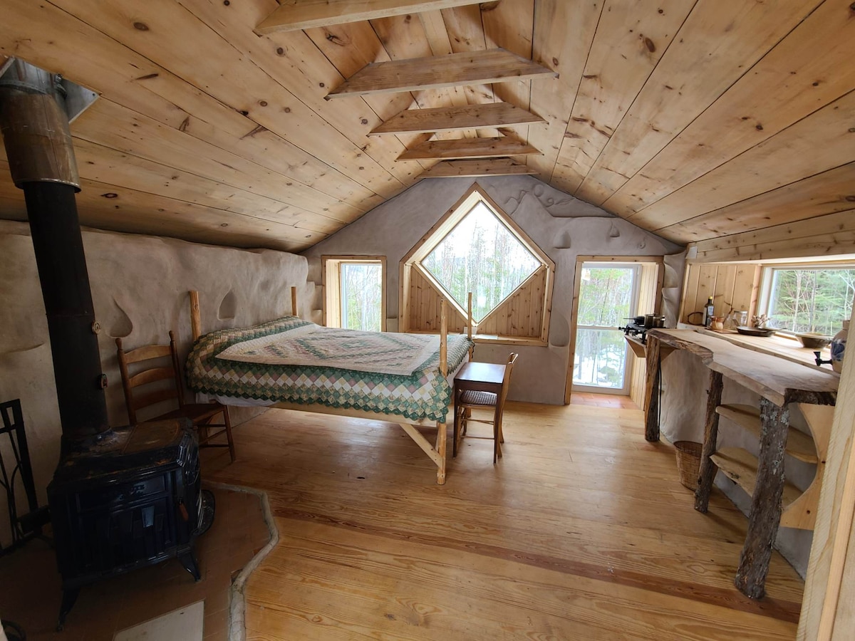 An open interior showcases natural wood walls and flooring, with a spacious bed framed by a unique diamond-shaped window. A wood stove is positioned nearby, while a small kitchen area features rustic shelving and a countertop made from natural materials.