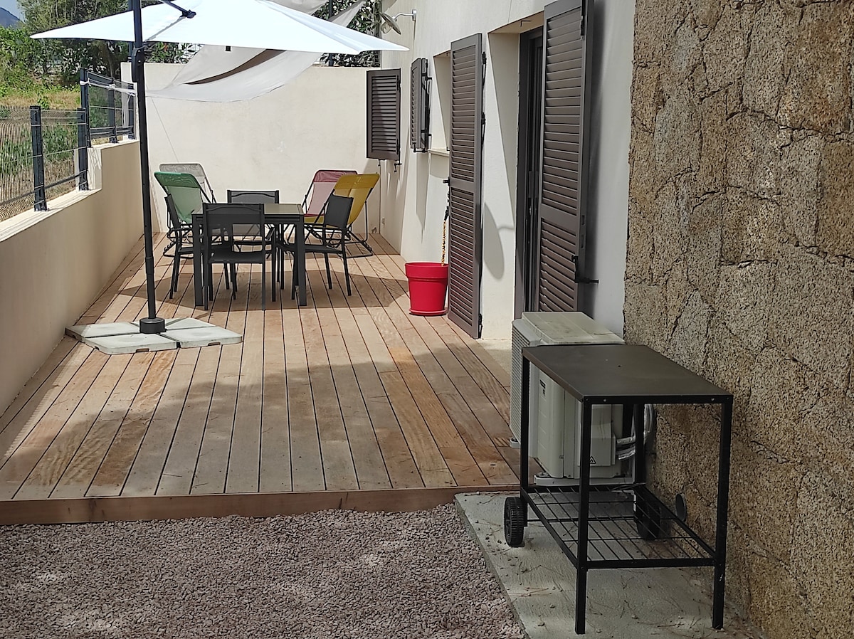 A large wooden terrace features a dining table with colorful chairs beneath a sunshade. An air conditioning unit is positioned beside a metal table, while a red container adds a touch of color. The surrounding landscape includes textured stone walls and greenery.