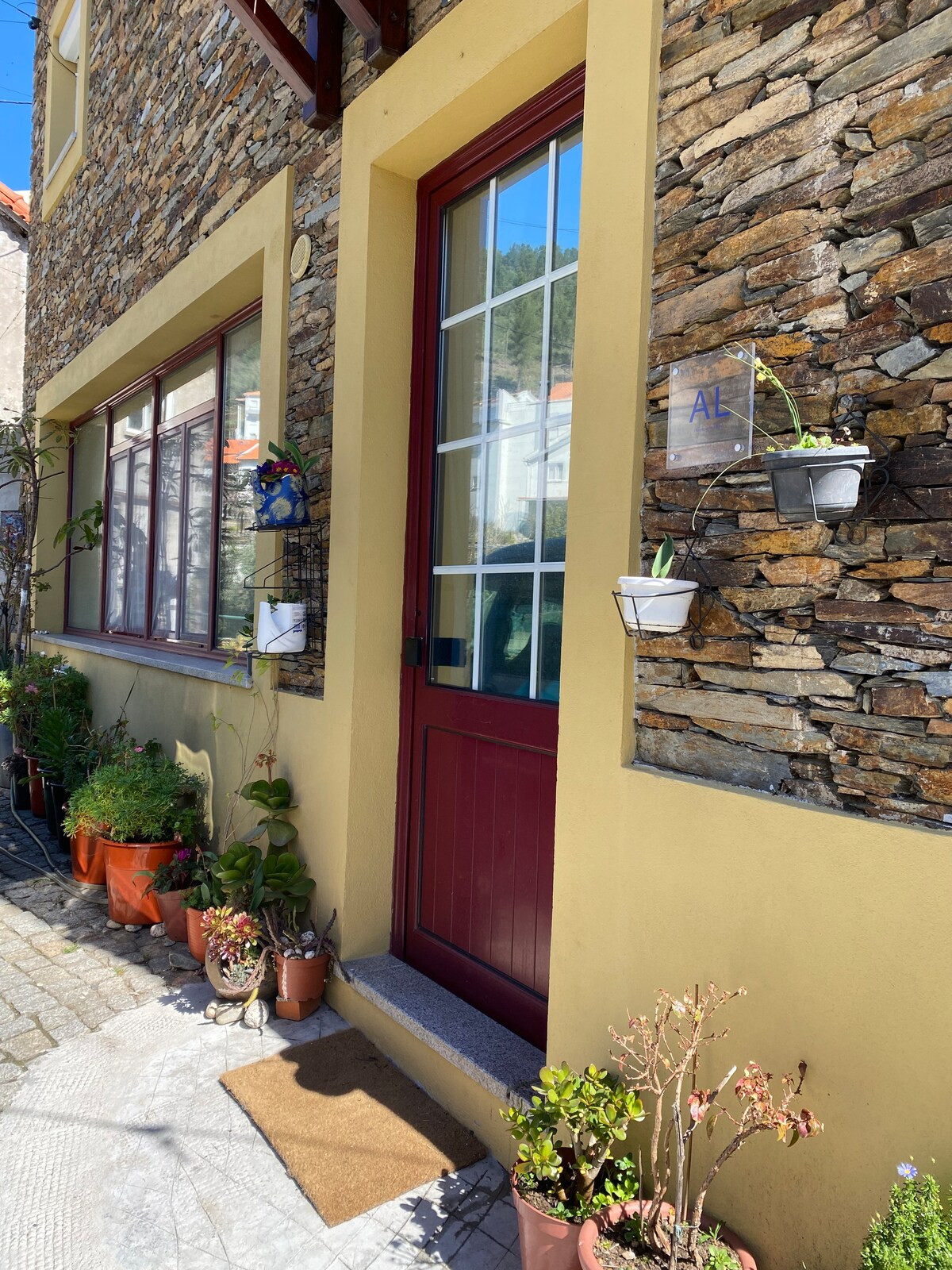 Entrance to the apartment is framed by a stone facade, featuring a maroon door with glass panels. Potted plants are arranged along the entryway, enhancing the welcoming feel while providing a glimpse into the serene outdoor space.