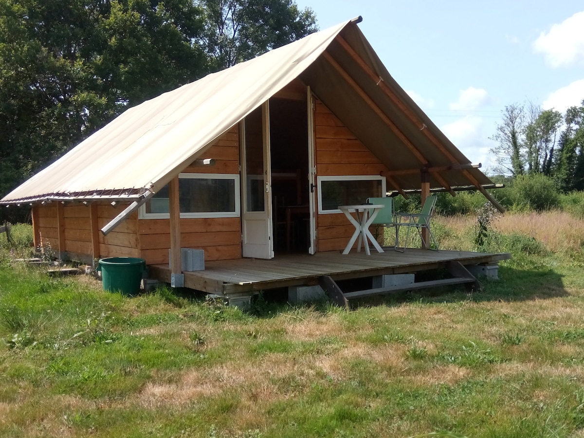 The cabin features a wooden exterior with a slanted canvas roof, situated on a raised deck. Large windows are visible on the front, providing natural light. Outside, a small table and chair set creates a space for outdoor relaxation. Lush greenery surrounds the cabin.