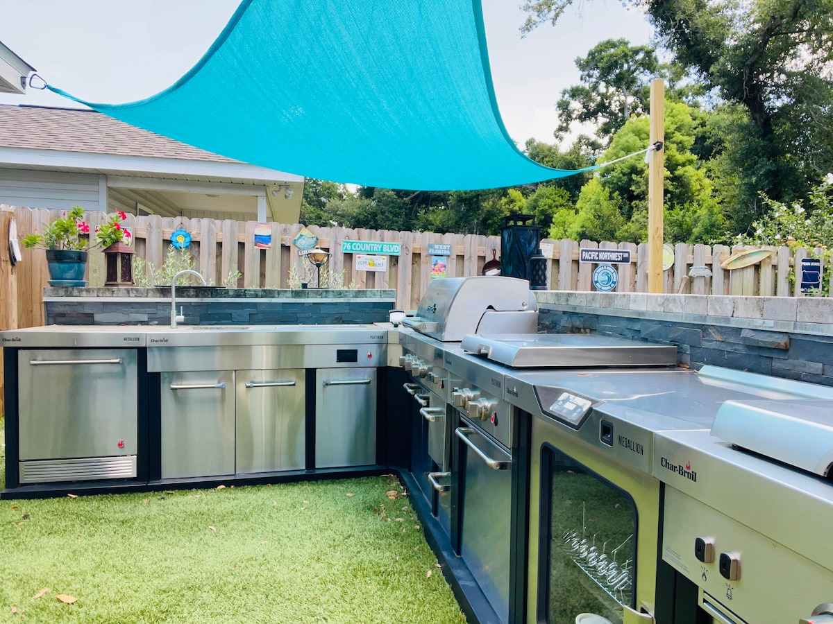 An outdoor kitchen is showcased with stainless steel appliances, including a grill and sink. A blue shade sail provides shelter from the sun. The kitchen area is surrounded by a stone countertop and features a grassy floor. Decorative signage and flowers enhance the space.