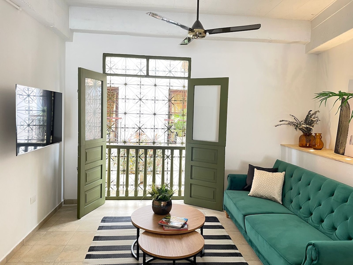 A bright living area is presented, featuring a green sofa and a round coffee table on a striped rug. Open doors reveal a view of the balcony with decorative wrought iron accents. A wall-mounted TV is positioned across from the seating, enhancing the space's comfort.
