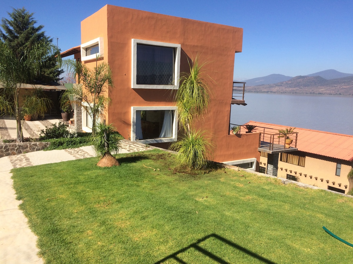 A two-story orange house is positioned near a tranquil body of water, framed by lush greenery. Large windows offer scenic views, and a spacious lawn leads to a stone pathway that enhances the outdoor space.