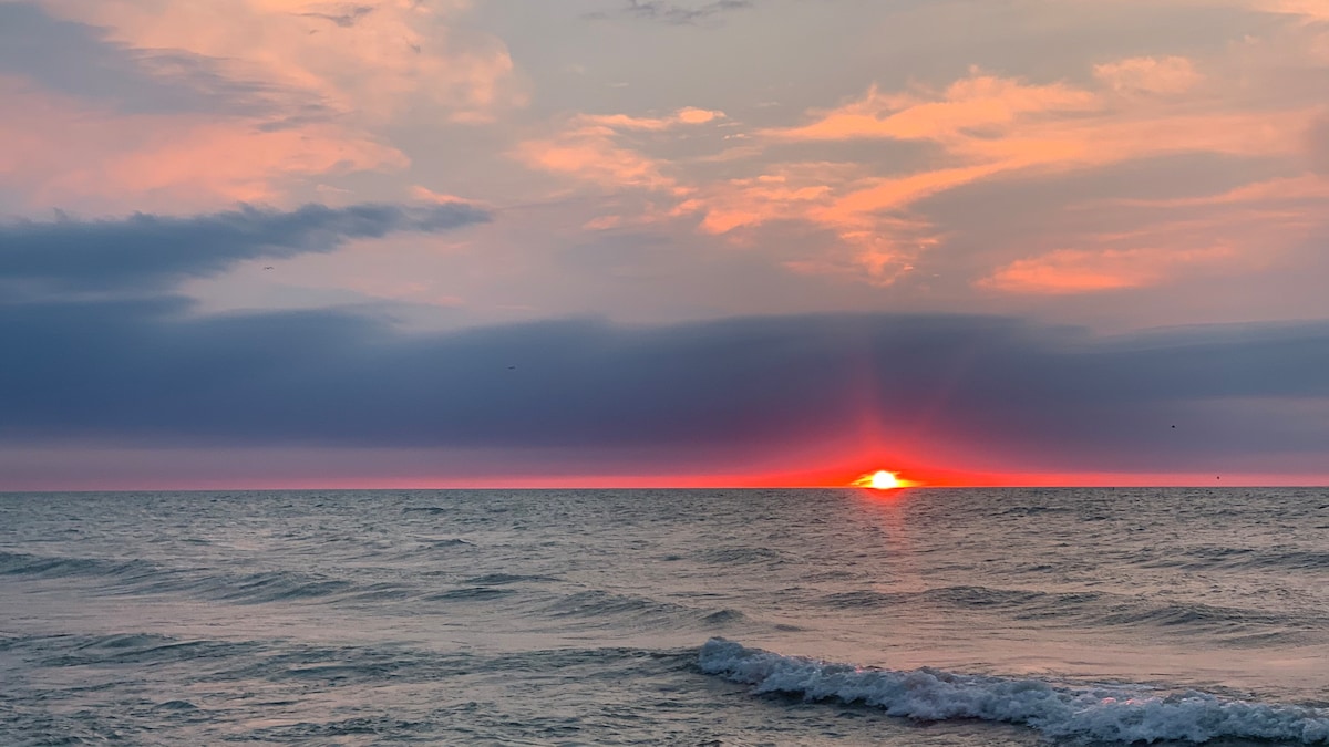 A serene view of Lake Michigan showcases the sun setting on the horizon, casting warm orange and pink hues across the sky. Gentle waves lap against the shore, reflecting the colors of dusk. The expansive water stretches toward the sky, creating a peaceful atmosphere.