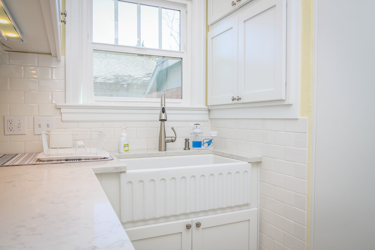 A bright kitchen space features a modern farmhouse sink set against white cabinetry and tiled backsplash. Natural light enters through a window above the sink, illuminating the clean countertop and minimalist design, creating a functional cooking area.