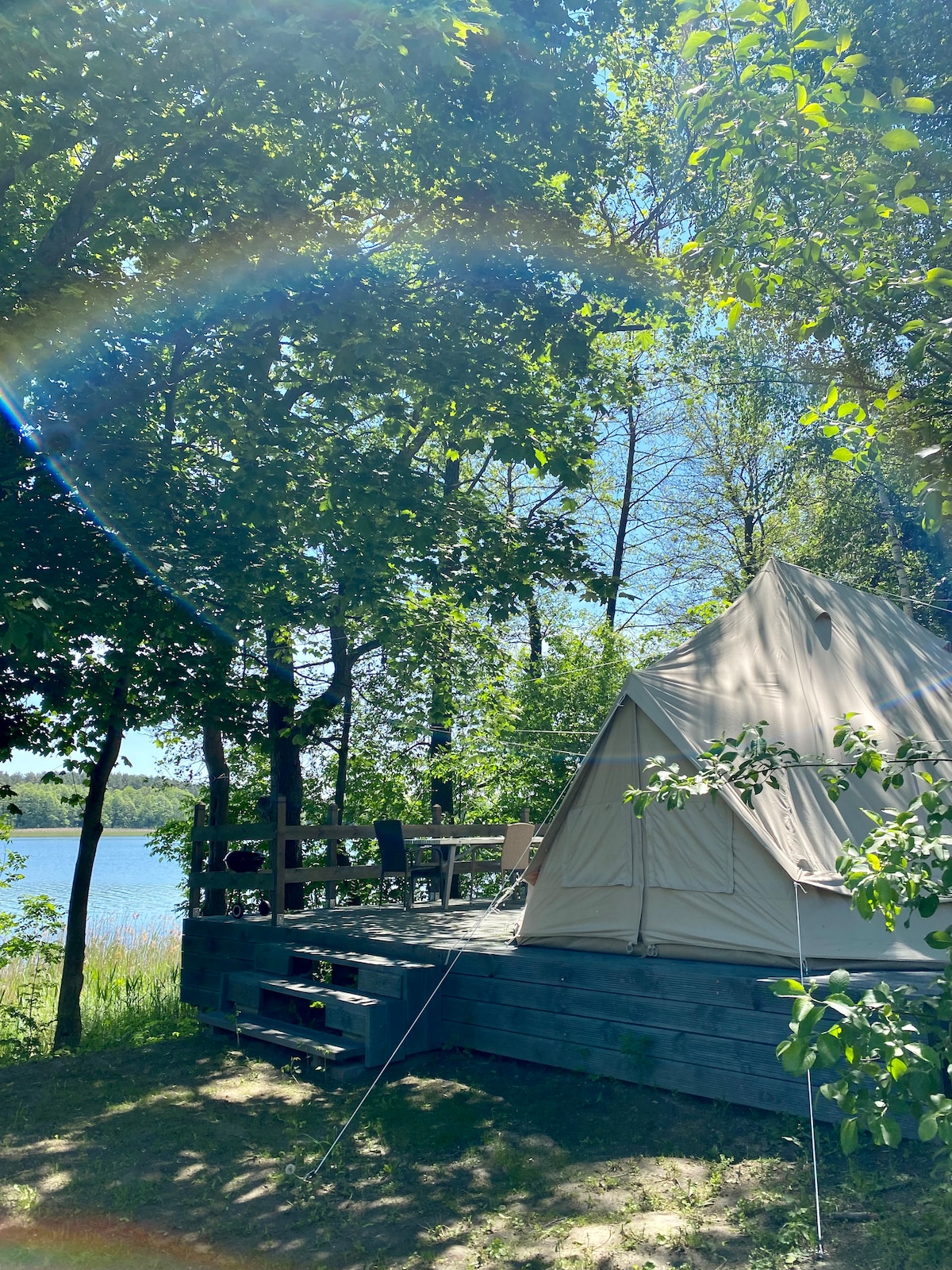 A spacious tent is nestled among trees, presenting a calm view of the nearby lake. A wooden deck is accessible, featuring outdoor furniture for relaxation. Sunlight filters through the leaves, enhancing the natural surroundings that envelop the area.