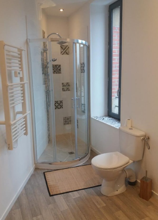 The bathroom features a glass shower enclosure with elegant tile work and a modern toilet. A towel warmer is mounted on the wall, and natural light enters through a tall window. A beige mat lies on the floor, adding warmth to the space.