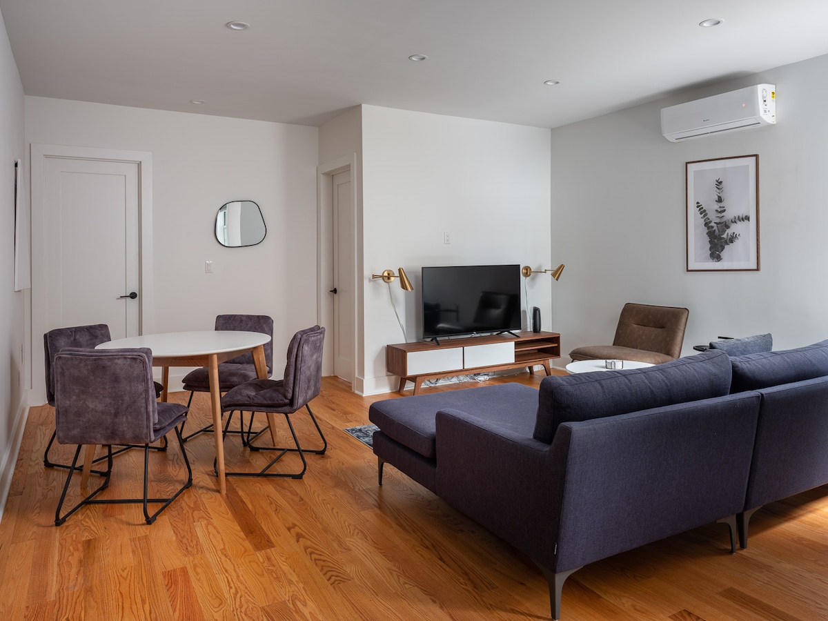 A modern living area features a comfortable dark sofa and contemporary chairs surrounding a circular dining table. A flat-screen TV is mounted on a sleek media console. Natural light fills the space through nearby windows, enhancing the light wood flooring.