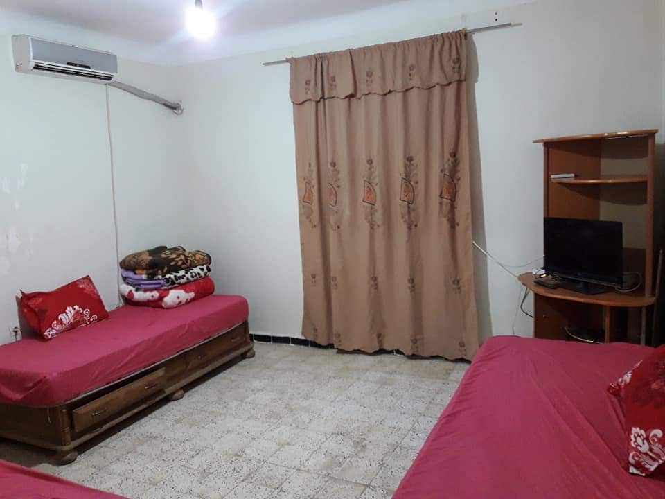 A simple and functional room features two beds with red bedding, placed against opposite walls. A curtain covers a window, while a wooden shelf supports a television in one corner. A stack of blankets rests on a nearby bed, adding a practical touch to the space.