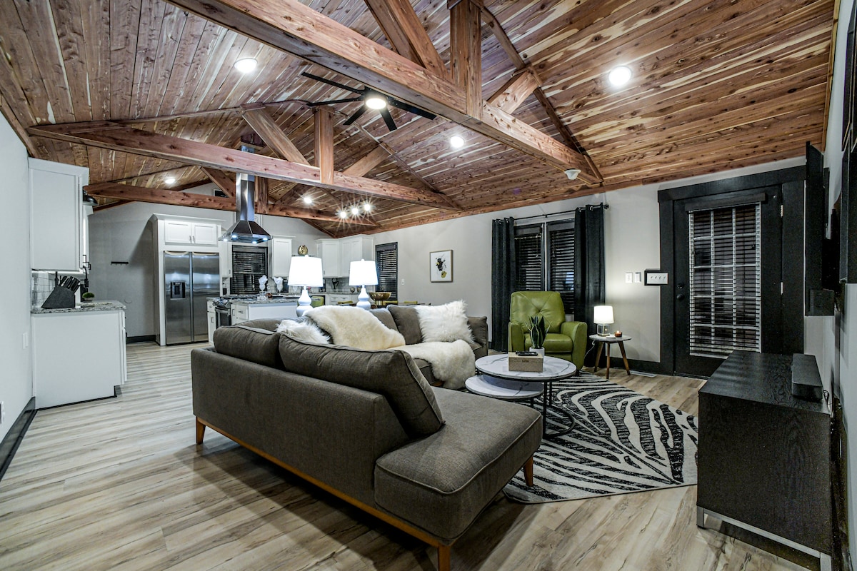 A spacious living area features high wooden beams and large windows allowing natural light. Comfortable seating is provided by a dark sofa and bright green chair. A round coffee table sits atop a stylish patterned rug, enhancing the inviting, open atmosphere. The kitchen is visible in the background.