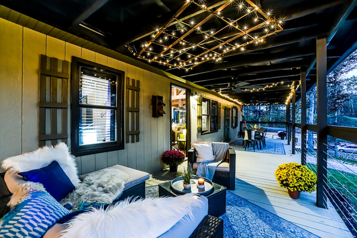A covered outdoor deck is illuminated by string lights, creating a cozy atmosphere. Seating includes a plush lounge chair and coffee table, accompanied by a potted plant. A dining area is visible in the background, surrounded by open space and natural views.