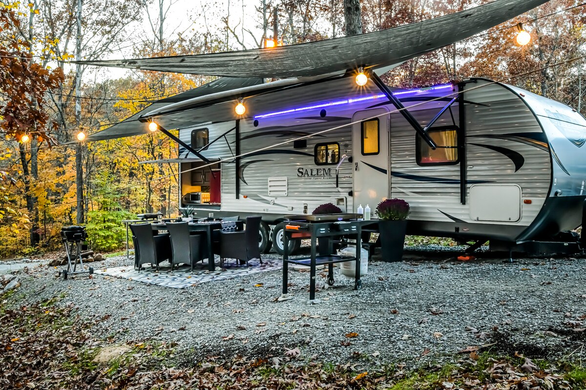 An outdoor seating area is set up beside a well-equipped camper, complete with a gas grill and a rugged blackstone. Warm string lights cast a cozy glow, with a shaded area providing relief from the sun, surrounded by autumn foliage.
