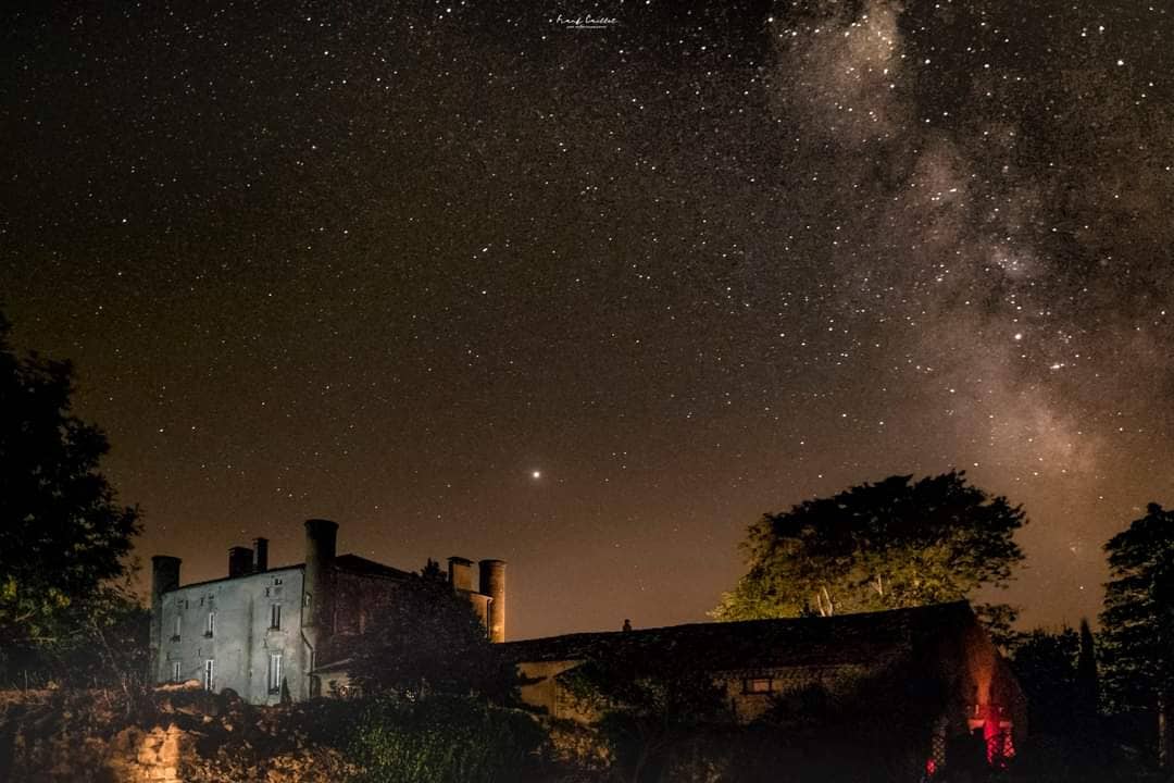 A historic building emerges against a clear night sky, revealing a cascade of stars with the Milky Way stretching overhead. The structure reflects soft lighting, and surrounding trees are silhouetted, enhancing the tranquil ambiance of the setting.
