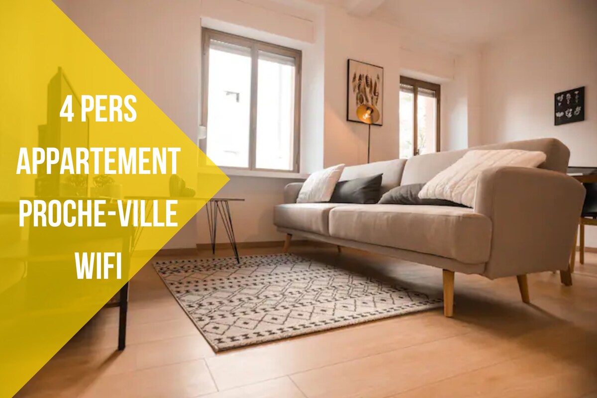 A bright living area features a spacious sofa with decorative cushions, positioned beside two windows that allow natural light to fill the room. A patterned rug lies on the wooden floor, complemented by a small side table for added convenience.