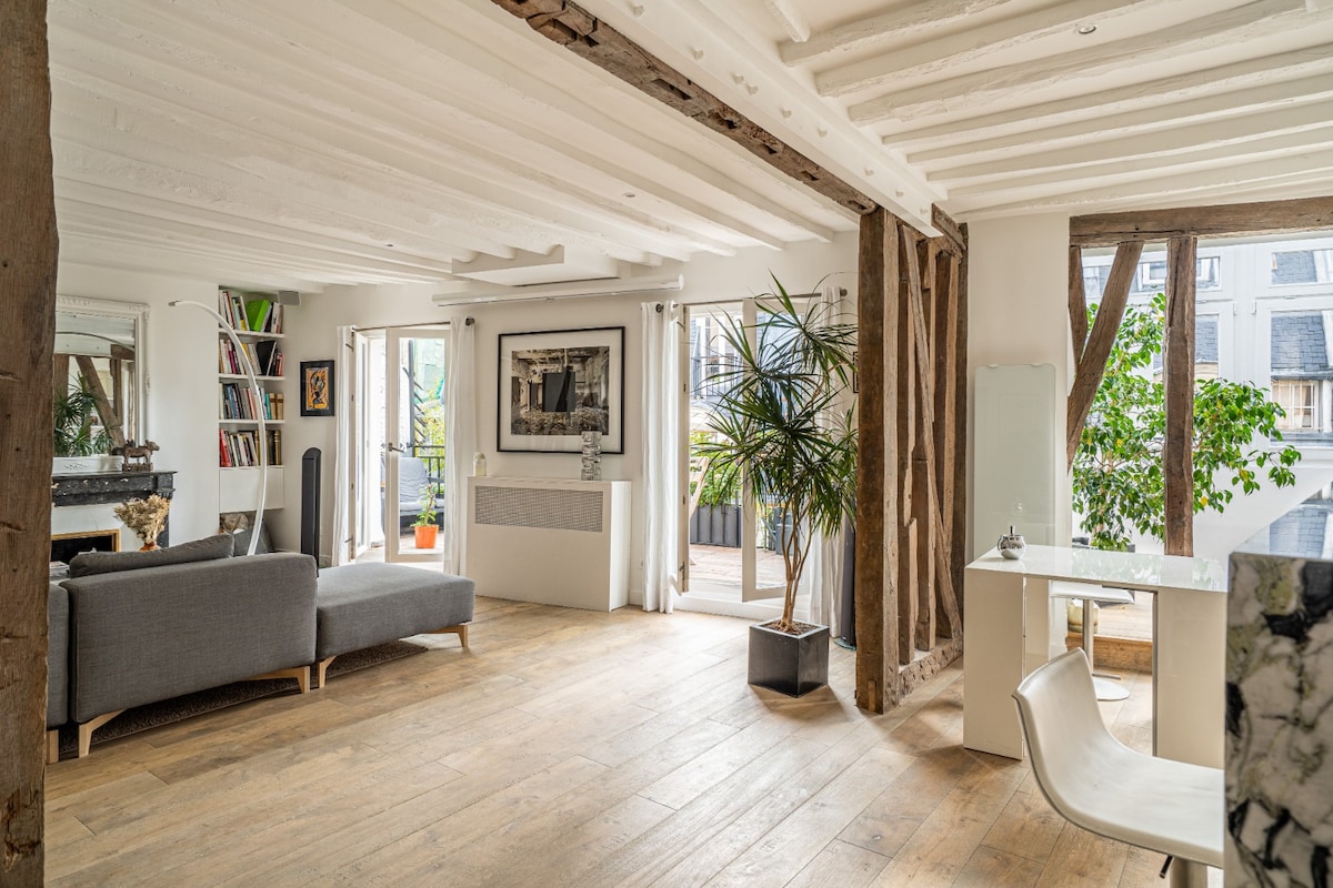 A bright and airy living space features wooden beams and a warm, light hardwood floor. A comfortable gray sofa is positioned near large glass doors that lead to a terrace. A simple dining table and chair are placed thoughtfully in the room, complemented by greenery from potted plants.
