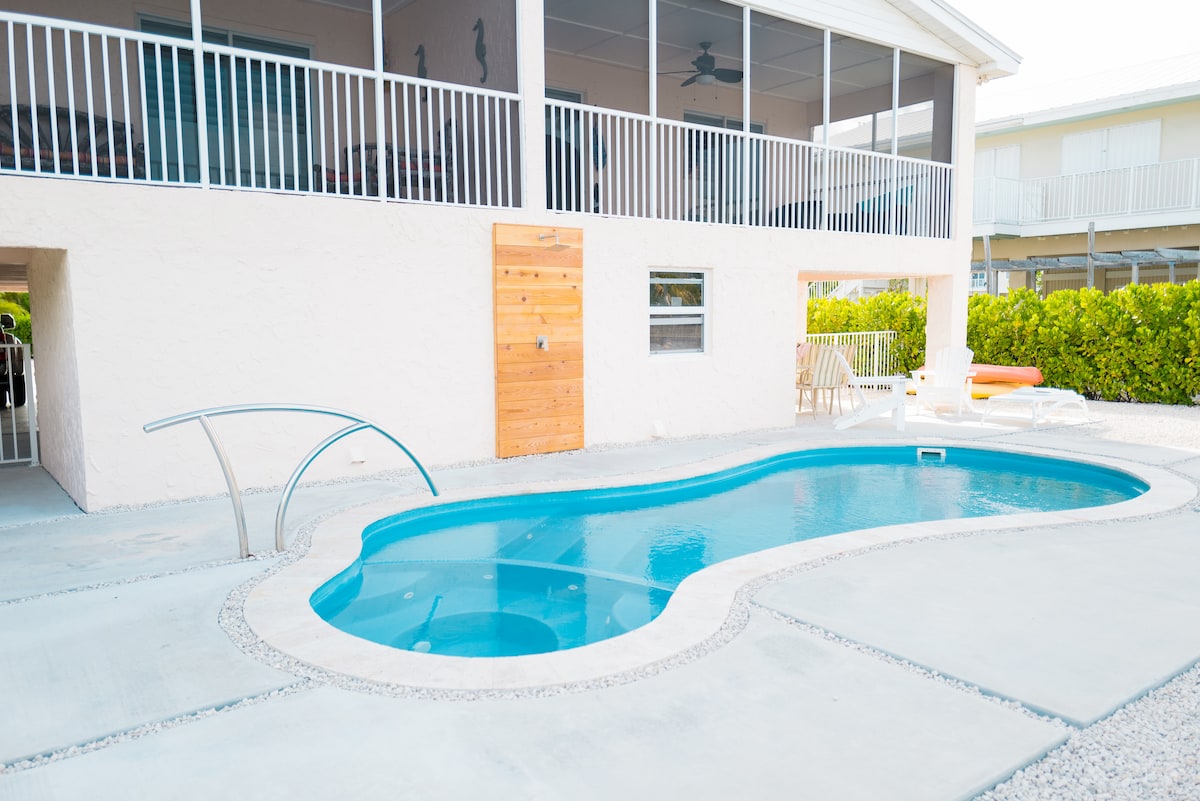 The saltwater pool features a gently curved shape and is surrounded by a spacious concrete patio. An outdoor shower is visible nearby, alongside lounge chairs and a dining area. The lower level of the house provides access to the pool area through a wooden door.