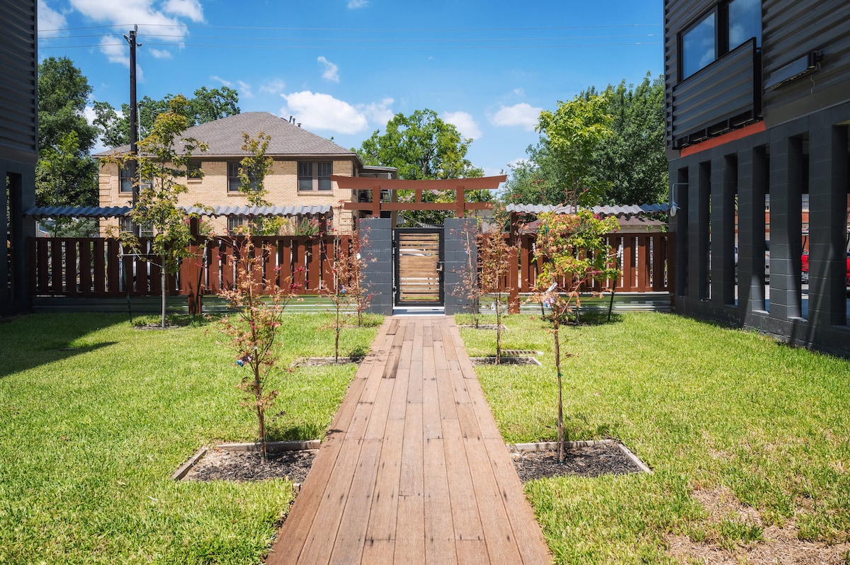 A landscaped outdoor area is visible, featuring a wooden pathway leading through neatly arranged greenery and small trees. A gate can be seen at the far end, framed by a wooden structure, enhancing the inviting atmosphere of the space.