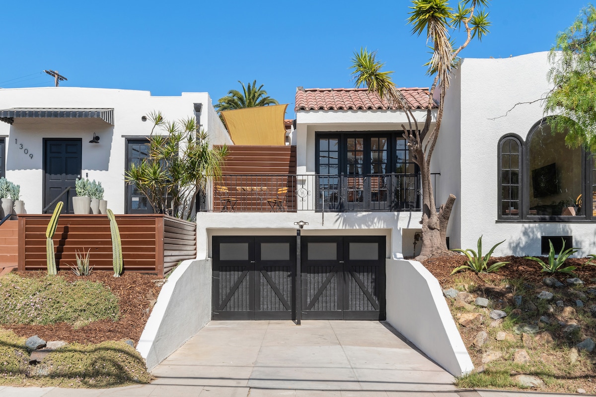 The exterior view features two distinct white homes with vibrant landscape elements, including palm trees and cacti. A spacious driveway with a double garage door is visible, providing access to the properties. A balcony overlooks the front garden, enhancing the outdoor space.