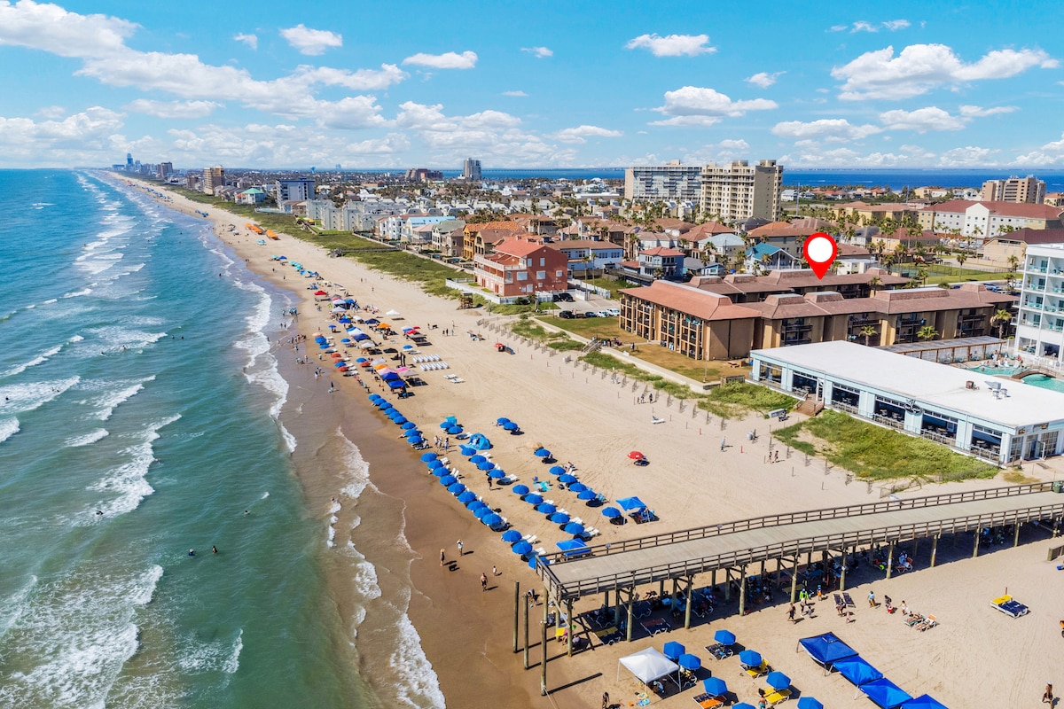 An aerial view captures the sandy beach lined with colorful umbrellas and sunbathers. Nearby, beachside establishments are visible, along with a boardwalk leading toward the water. The coastal landscape showcases the Gulf of Mexico under a clear blue sky.