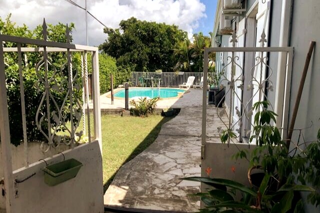 A gated entrance opens to a paved path leading to a shared pool area. Lush greenery surrounds the space, providing a sense of privacy. The pool is visible in the background, framed by an enclosed deck. Bright sunlight casts soft shadows on the ground.