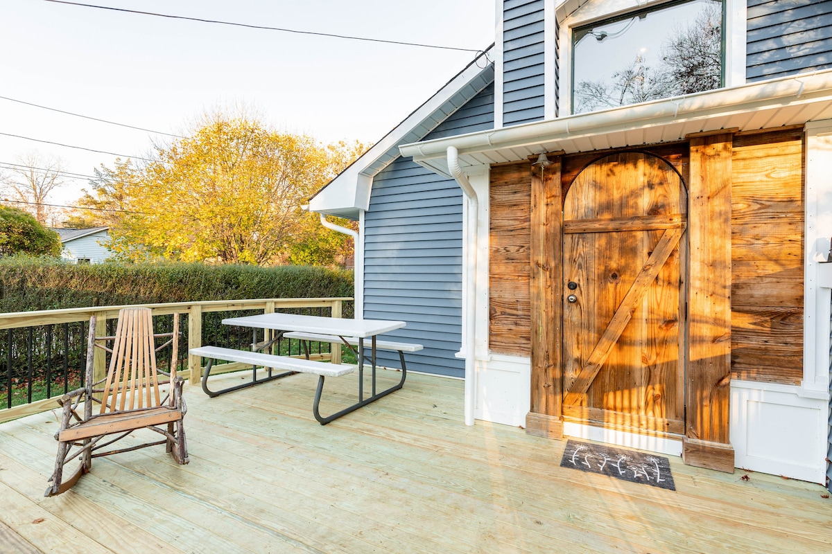 A rustic wooden door stands prominently on the facade of the blue house, inviting entry. Beside it, a stylish picnic table is positioned on the sunlit deck, complemented by a classic wooden rocking chair. Lush greenery surrounds the space, enhancing the natural setting.