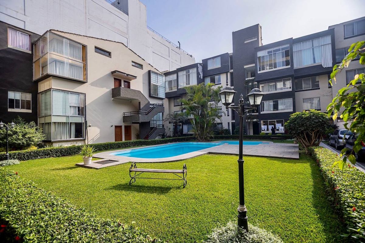 A serene courtyard features a well-maintained lawn, bordered by shrubbery. A swimming pool is centrally located, flanked by steps leading to the surrounding apartments. Charming lantern-style lighting adds a decorative touch, and tropical plants enhance the tranquil ambiance.
