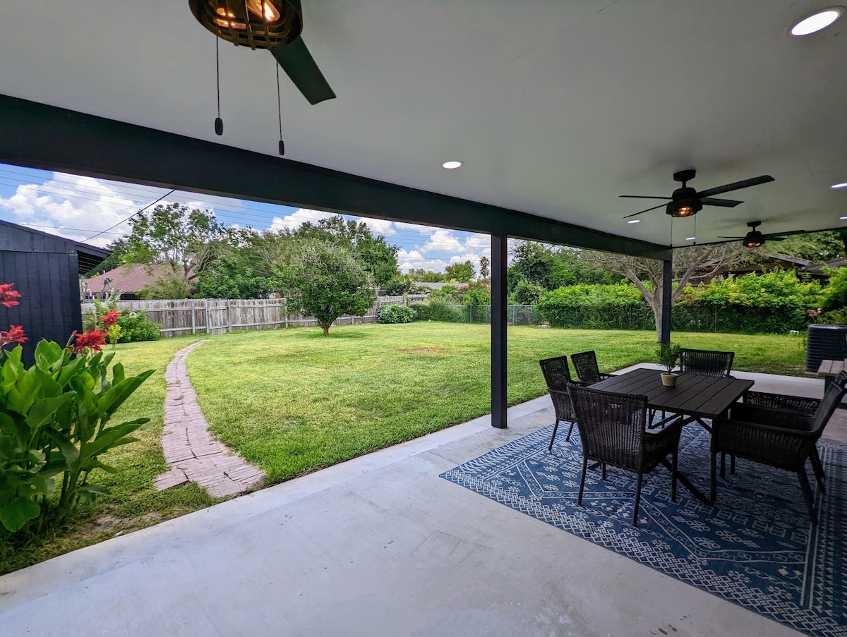 A large covered patio area provides ample seating with a table surrounded by woven chairs. Fans are installed overhead, and vibrant green landscaping fills the backyard, featuring a pathway winding through the grass and mature trees in the distance.