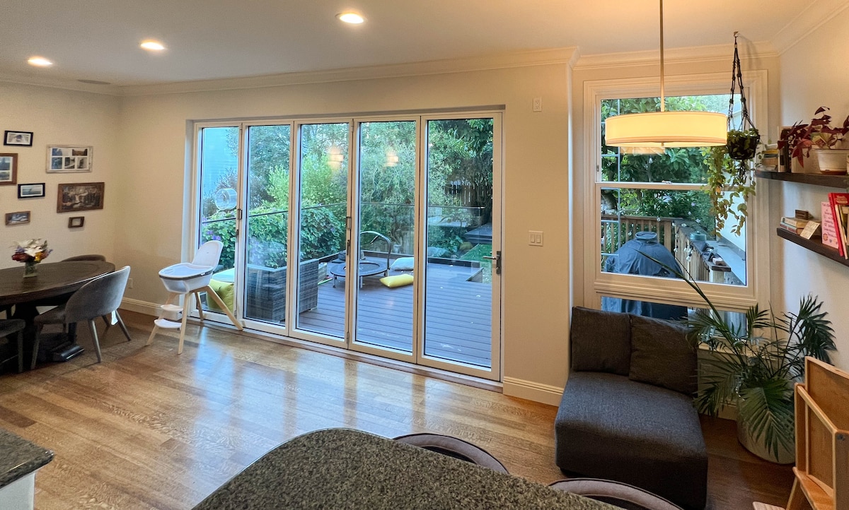 A spacious living area is highlighted by large glass doors that open to a patio and garden area, allowing natural light to fill the room. Comfortable seating is arranged around a round dining table, with a children's high chair positioned nearby.