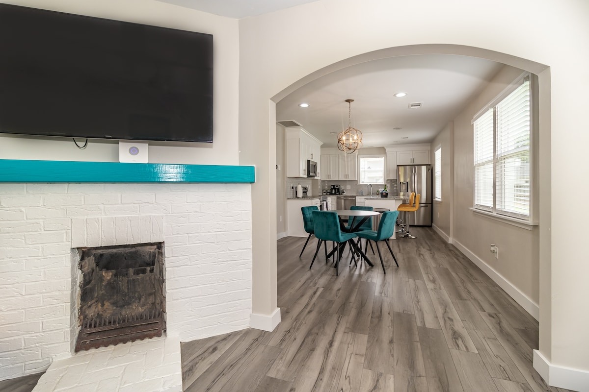 A modern living space features a large flat-screen TV mounted above a white brick fireplace. An archway leads to a dining area with a round table surrounded by stylish chairs. Ample natural light is provided by large windows adjacent to a well-equipped kitchen.